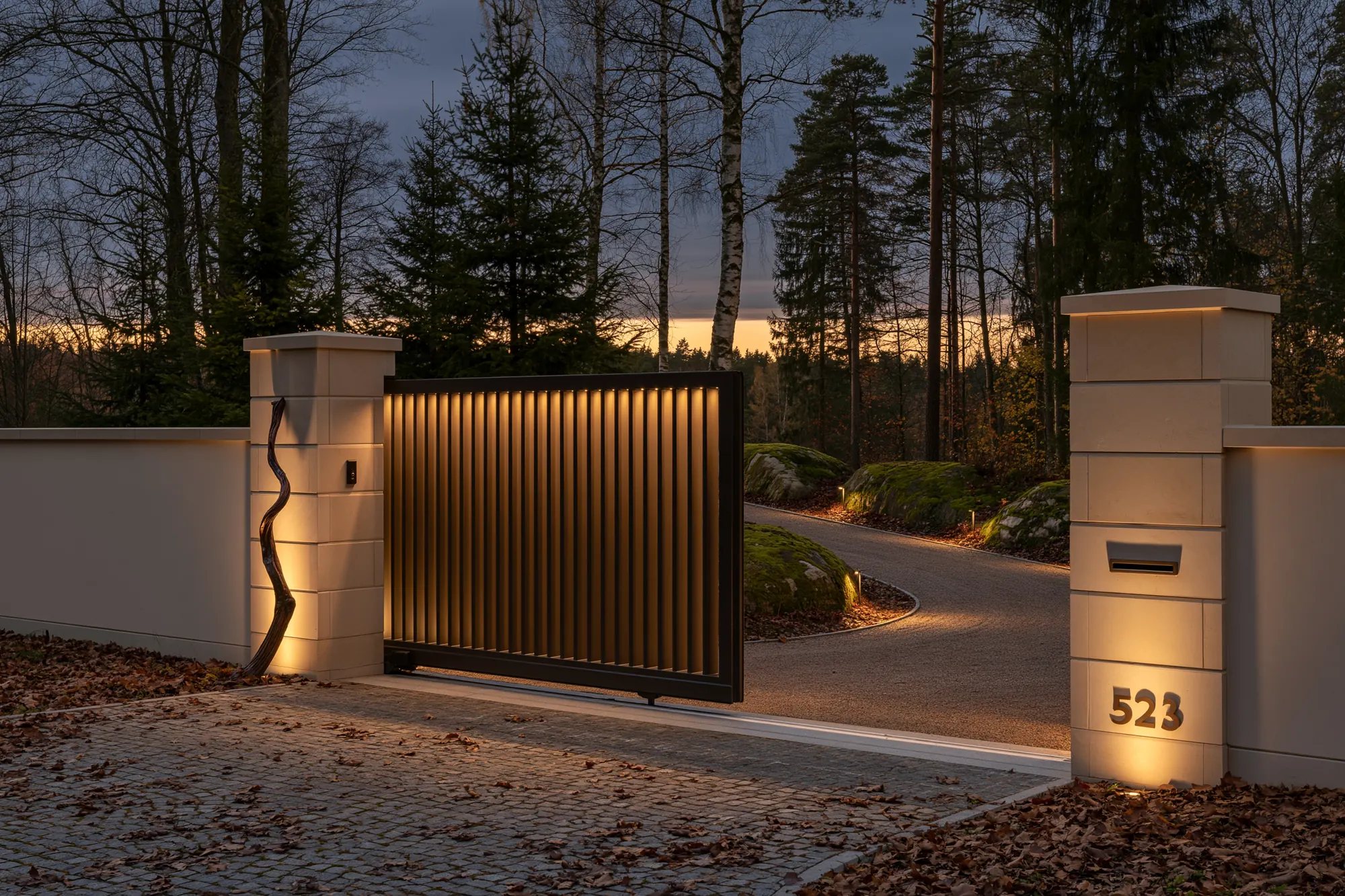 08-gate A slatted timber gate sliding open between pale stone piers at dusk, a gravel drive curving into the pines.